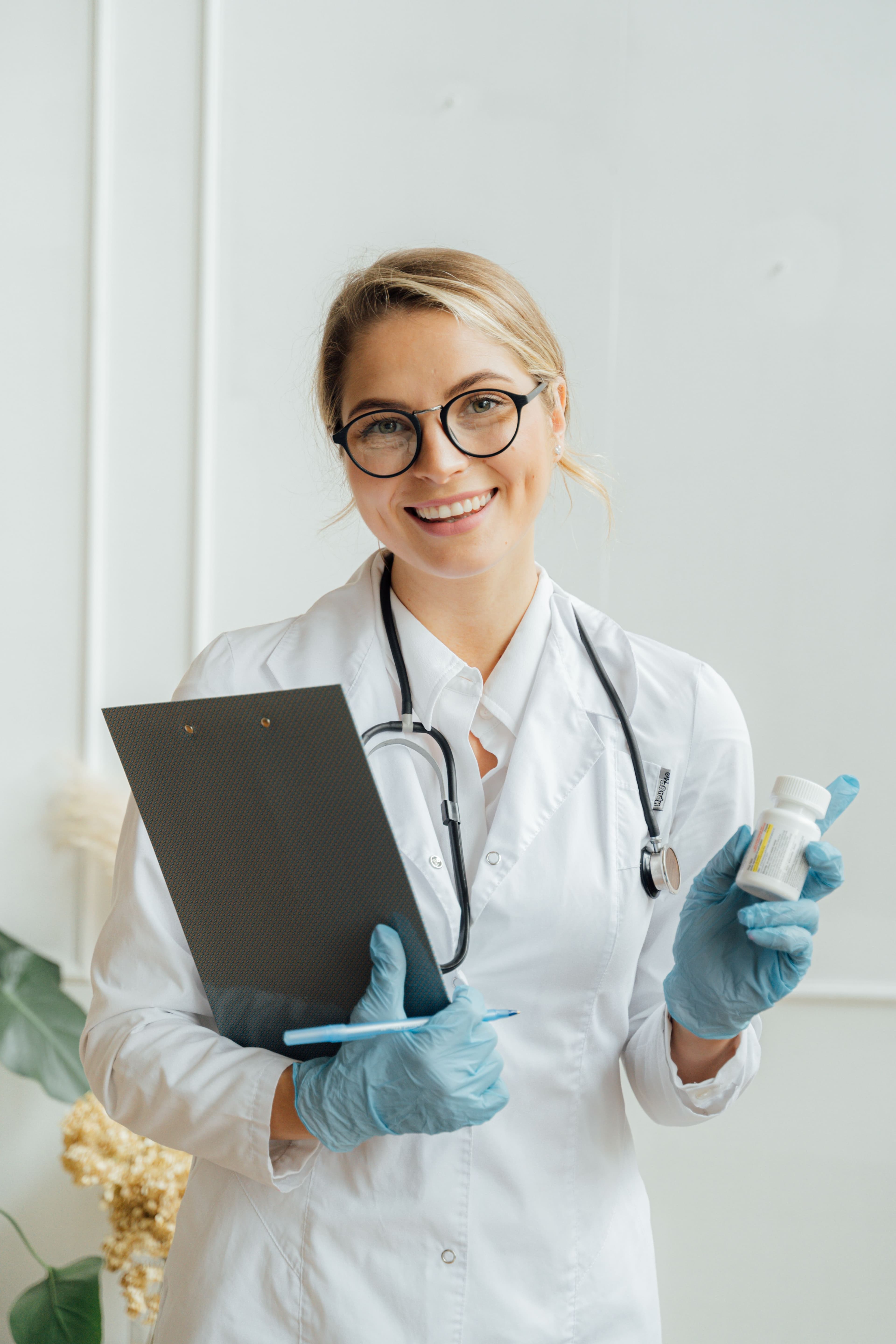 A smiling doctor in a white coat holding a clipboard and a bottle of medication