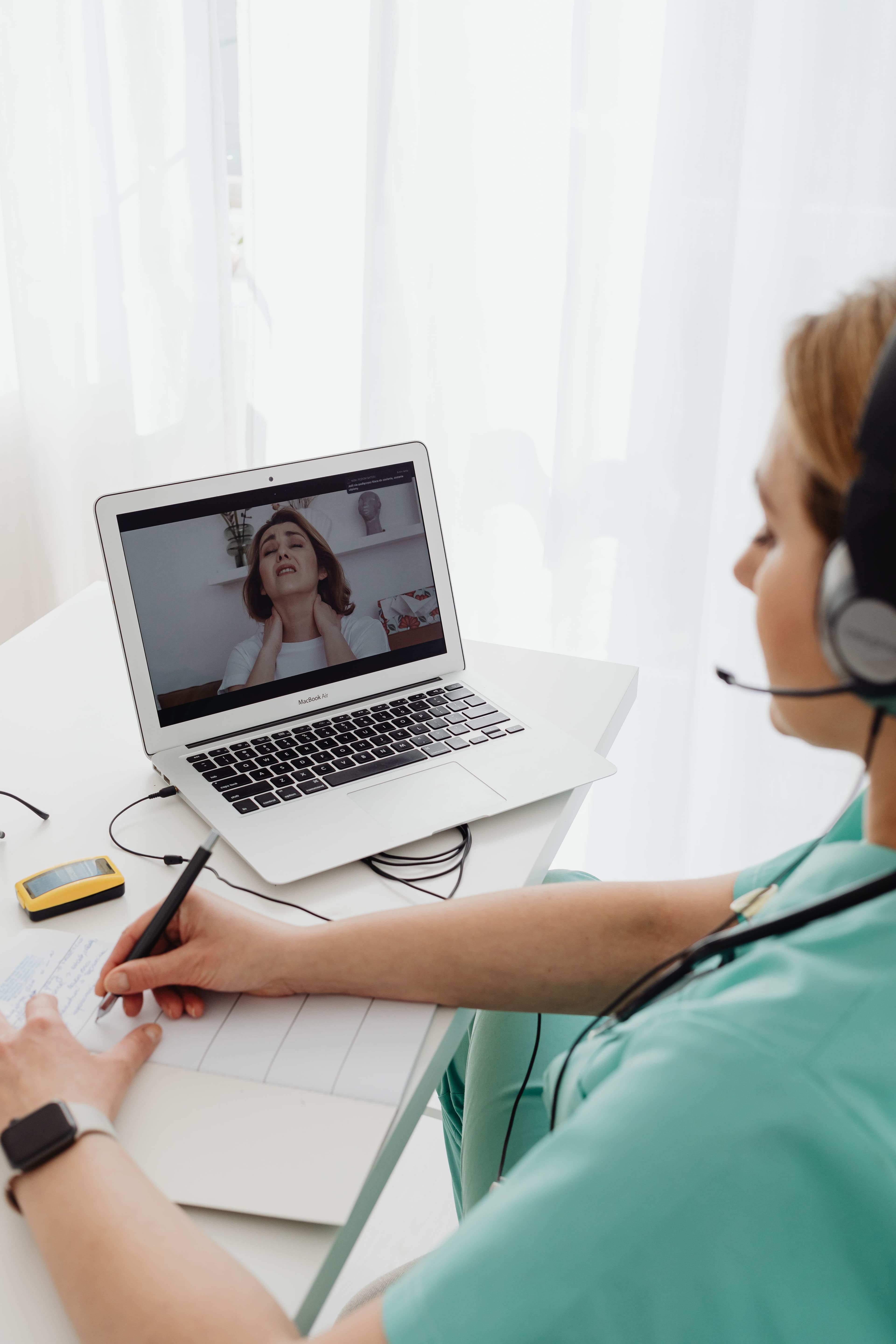A clinician wearing a headset takes notes during a video consultation with a patient on her laptop